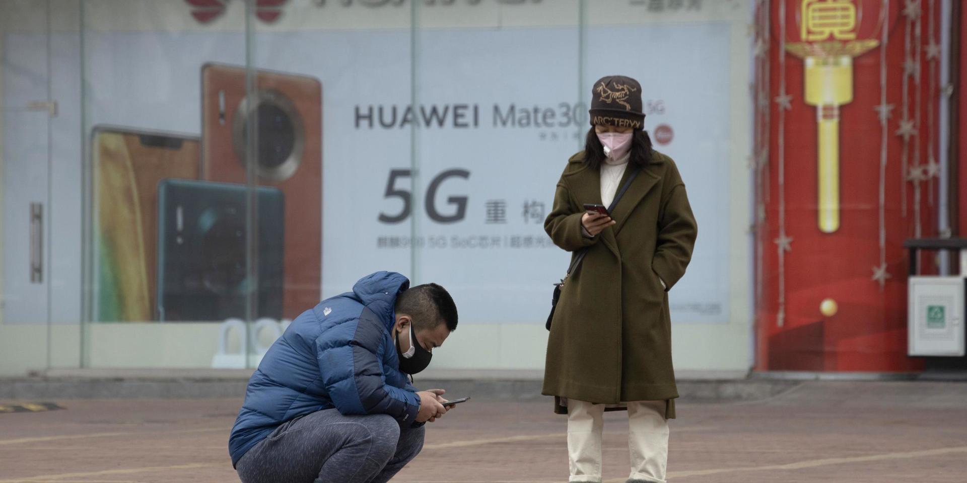 In this Sunday, March 8, 2020, photo, residents wearing masks look at their smartphones near an advertisement for 5G smartphones from Chinese tech giant Huawei in Beijing. Factories in China, struggling to reopen after the coronavirus shut down the economy, face a new threat from U.S. anti-disease controls that might disrupt the flow of microchips and other components they need. For most people, the new coronavirus causes only mild or moderate symptoms. For some it can cause more severe illness. (AP Photo/Ng Han Guan)  XHG102